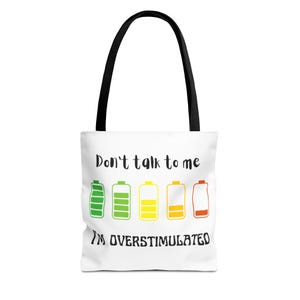 Può includere: Borsa tote bianca con manici neri. Presenta la scritta "Don't talk to me, I'm overstimulated" e icone di batteria verdi, gialle e rosse, che rappresentano i livelli di energia.