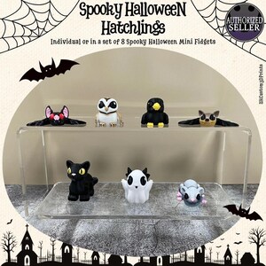 May include: A clear display shelf holds eight miniature Halloween-themed figurines. The figurines include a black cat, a ghost, a bat, an owl, a bird, and a mouse. The text "Spooky Halloween Hatchlings" is at the top of the image.