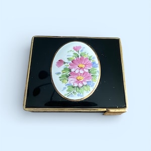 May include: A black compact with a gold-colored trim. The compact features an oval floral design with pink and yellow flowers on a white background. The flowers are surrounded by green leaves and blue accents.