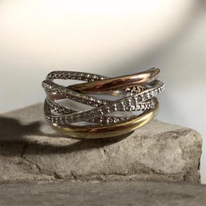 May include: A multi-tone ring featuring a crisscross design with three distinct bands. One band is smooth yellow gold, one is smooth rose gold, and the third is a silver-toned band encrusted with small, clear, sparkling stones throughout.