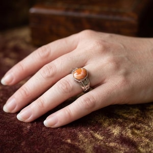 May include: A silver ring featuring a smooth, oval-shaped orange stone with white marbling. The band of the ring has a wide, metallic design that tapers toward the back, securely holding the polished cabochon stone in a simple metal setting.