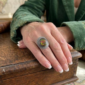 May include: A silver ring featuring an oval-shaped, iridescent stone with swirling green, red, and orange hues. The stone is set within a textured, braided silver frame, creating a detailed border around the central gemstone.
