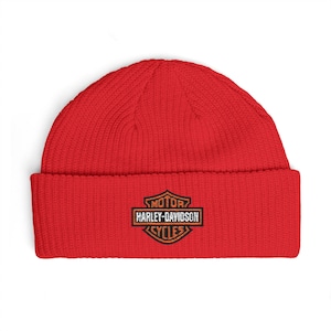 May include: A bright red beanie hat with a ribbed knit texture. The hat features the Harley-Davidson Cycles logo embroidered on the front in black, orange, and white. The logo is rectangular with the brand name and the word "cycles" below.