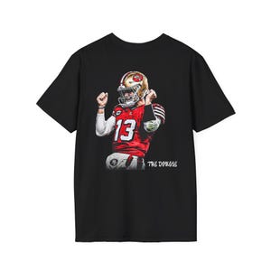 May include: Black t-shirt featuring a graphic of a football player in a red and gold uniform, with the number 13. The text "THE DOUGIE" is printed below the image. The player is wearing a helmet and raising his arms in a celebratory pose.