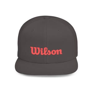 Op de afbeelding: Een donkergrijze baseballcap met een platte klep. Het woord "Wilson" is in koraalrood op de voorkant geborduurd. De cap heeft een gestructureerde pasvorm en een klassiek, sportief ontwerp.