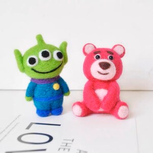 Könnte beinhalten: Zwei Filzspielzeuge, ein grüner Außerirdischer mit drei Augen und ein rosa Bär mit brauner Nase, die auf einer weißen Oberfläche sitzen.