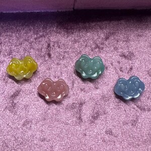 May include: Four small, colorful, resin smiley face charms. The charms are shaped like a cloud and have a glossy finish. The colors are yellow, pink, light blue, and dark blue.