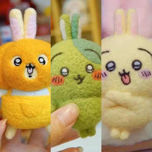 Könnte beinhalten: Drei handgefertigte Hasenfiguren aus Filz in Gelb, Grün und Hellgelb. Jeder Hase hat einen einzigartigen Ausdruck, mit Details wie rosa Wangen und unterschiedlichen Ohrfarben. Die Hasen sind mit cartoonartigen Merkmalen gestaltet.