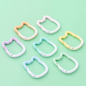 Puede incluir: Seis clips metálicos con forma de gato de colores vivos con un diseño degradado. Los clips están dispuestos en círculo sobre un fondo verde.