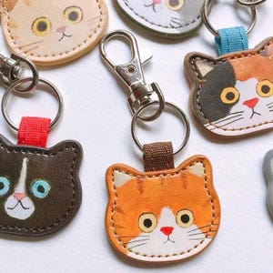May include: A selection of cat-shaped keyrings in shades of brown, orange, and grey. Each keyring has a metal ring and clasp. The cat faces are embroidered with details like eyes, noses, and whiskers.