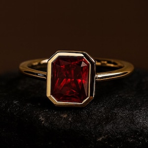 Pode incluir: Um anel de ouro com uma pedra preciosa vermelha escura retangular. A pedra é engastada em uma moldura de ouro, e a banda é de ouro polido simples. O anel é exibido em uma superfície escura e texturizada.