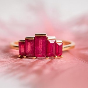 Pode incluir: Um anel de ouro com cinco pedras preciosas retangulares de um vermelho vibrante. As pedras estão dispostas em uma fileira, com a pedra central ligeiramente maior. O anel está sobre um fundo rosa suave, criando um contraste marcante.