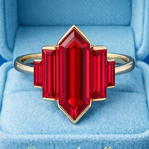 May include: A gold ring with a large, rectangular, red gemstone center and smaller side stones. The ring is set in a gold frame and sits in a blue velvet box. The text "Dreamsofjewellry" is at the bottom of the box.