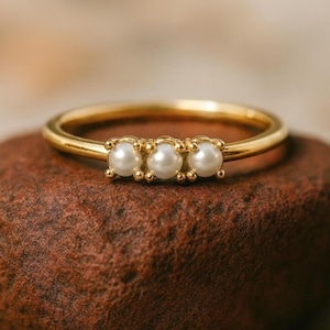 May include: A gold ring with three small, round, white pearls set in a row. The ring has a thin band and is displayed on a brown surface. The pearls are evenly spaced, adding a touch of elegance to the design.