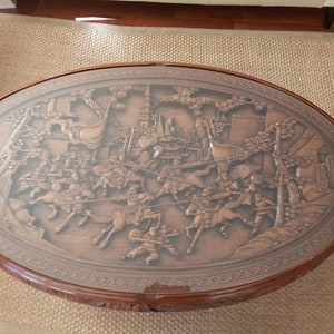 May include: A round wooden coffee table with a glass top. The table top features a carved design of a battle scene with figures on horseback.