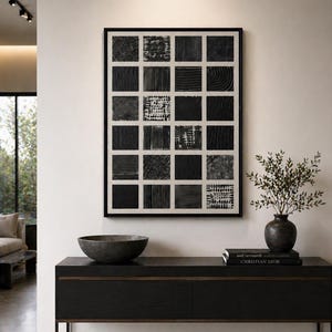 May include: A framed wall art piece featuring a grid of twenty-four black squares with varied textures on a white background. The artwork is displayed above a black console table holding a dark bowl, a vase with greenery, and two stacked books.