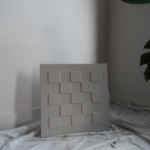 Pu&ograve; includere: Un'opera d'arte da parete quadrata in una tonalit&agrave; grigio chiaro neutra caratterizzata da un motivo a scacchiera in rilievo. Il design &egrave; composto da quadrati lisci alternati a quadrati con sottili texture a linee verticali, creando una composizione geometrica tattile.