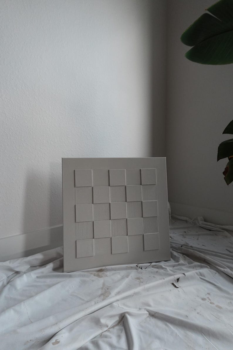 Pu&ograve; includere: Un'opera d'arte da parete quadrata in una tonalit&agrave; grigio chiaro neutra caratterizzata da un motivo a scacchiera in rilievo. Il design &egrave; composto da quadrati lisci alternati a quadrati con sottili texture a linee verticali, creando una composizione geometrica tattile.
