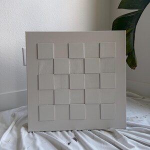 Pu&ograve; includere: Un'opera d'arte da parete quadrata di colore bianco sporco con un motivo a scacchiera in rilievo. Il design &egrave; composto da un'alternanza di quadrati lisci e quadrati con texture fini, lineari, verticali o orizzontali, creando una griglia tattile.
