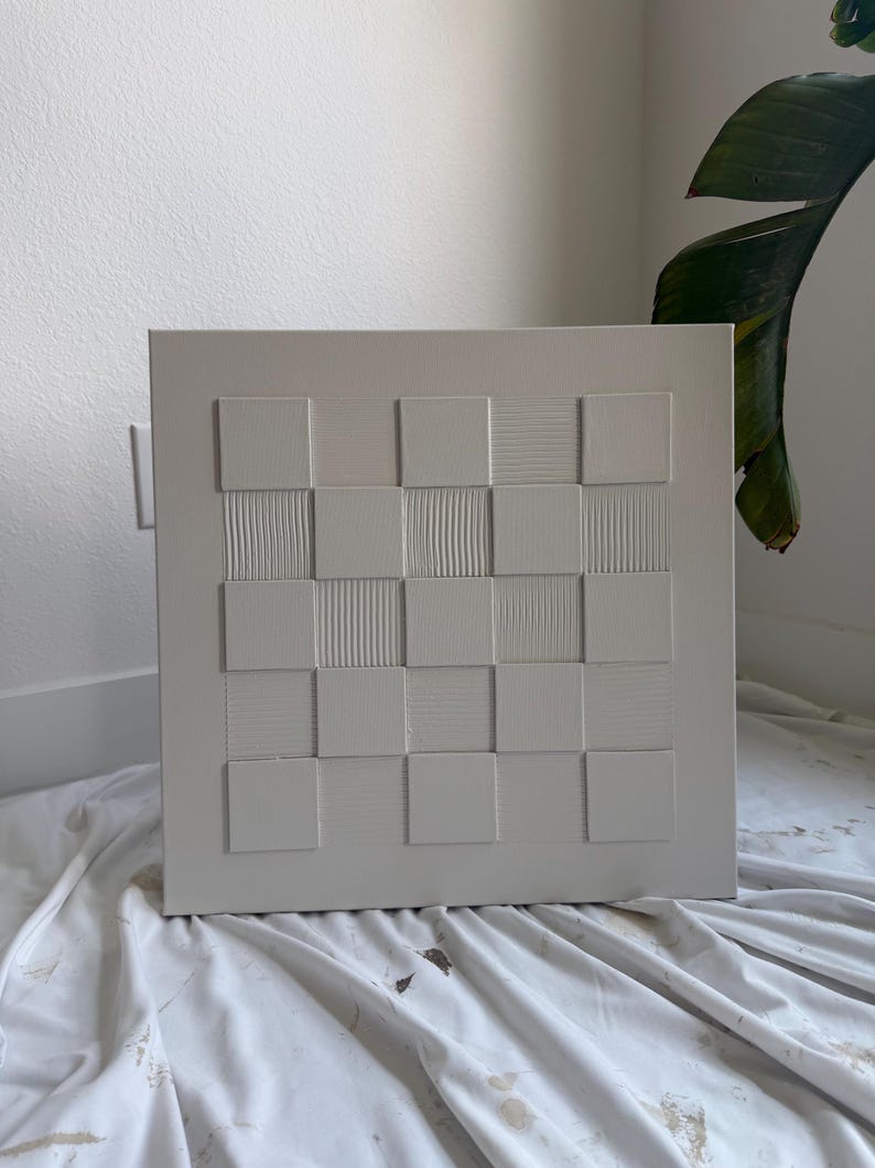 Pu&ograve; includere: Un'opera d'arte da parete quadrata di colore bianco sporco con un motivo a scacchiera in rilievo. Il design &egrave; composto da un'alternanza di quadrati lisci e quadrati con texture fini, lineari, verticali o orizzontali, creando una griglia tattile.