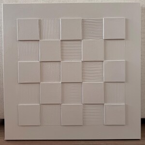 Pu&ograve; includere: Un'opera d'arte da parete quadrata di colore bianco caratterizzata da una griglia di quadrati in rilievo. Il design alterna superfici lisce e piatte a quadrati testurizzati con linee orizzontali o verticali, creando un motivo a rilievo monocromatico.