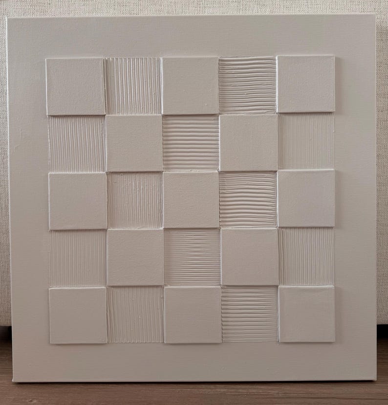 Pu&ograve; includere: Un'opera d'arte da parete quadrata di colore bianco caratterizzata da una griglia di quadrati in rilievo. Il design alterna superfici lisce e piatte a quadrati testurizzati con linee orizzontali o verticali, creando un motivo a rilievo monocromatico.