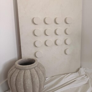 Peut inclure: Une &oelig;uvre d'art murale textur&eacute;e de ton neutre avec une grille d'&eacute;l&eacute;ments circulaires en relief. &Agrave; c&ocirc;t&eacute;, un grand vase en c&eacute;ramique gris-beige textur&eacute; avec des rainures verticales. Le vase a une large ouverture et repose sur un tissu blanc.