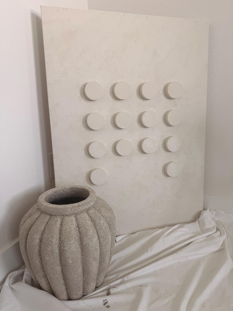 Peut inclure: Une &oelig;uvre d'art murale textur&eacute;e de ton neutre avec une grille d'&eacute;l&eacute;ments circulaires en relief. &Agrave; c&ocirc;t&eacute;, un grand vase en c&eacute;ramique gris-beige textur&eacute; avec des rainures verticales. Le vase a une large ouverture et repose sur un tissu blanc.