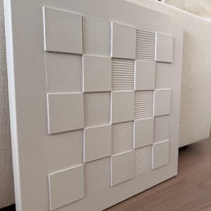 Pu&ograve; includere: Un'opera d'arte da parete quadrata di colore bianco caratterizzata da una griglia di quadrati in rilievo. Il design alterna quadrati lisci e piatti a quadrati con texture lineari verticali o orizzontali, creando un motivo geometrico monocromatico tattile.