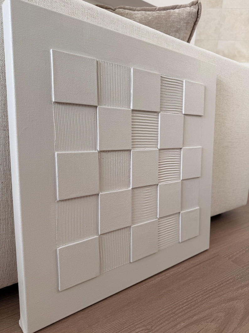 Pu&ograve; includere: Un'opera d'arte da parete quadrata di colore bianco caratterizzata da una griglia di quadrati in rilievo. Il design alterna quadrati lisci e piatti a quadrati con texture lineari verticali o orizzontali, creando un motivo geometrico monocromatico tattile.