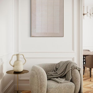 May include: A neutral-toned living room scene featuring a beige armchair with a gray knitted throw. A small side table holds a decorative vase. A framed abstract artwork hangs on the wall, adding a touch of modern art.