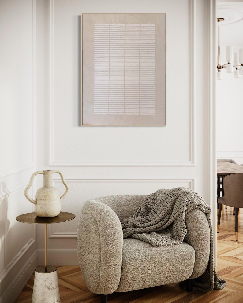 May include: A neutral-toned living room scene featuring a beige armchair with a gray knitted throw. A small side table holds a decorative vase. A framed abstract artwork hangs on the wall, adding a touch of modern art.