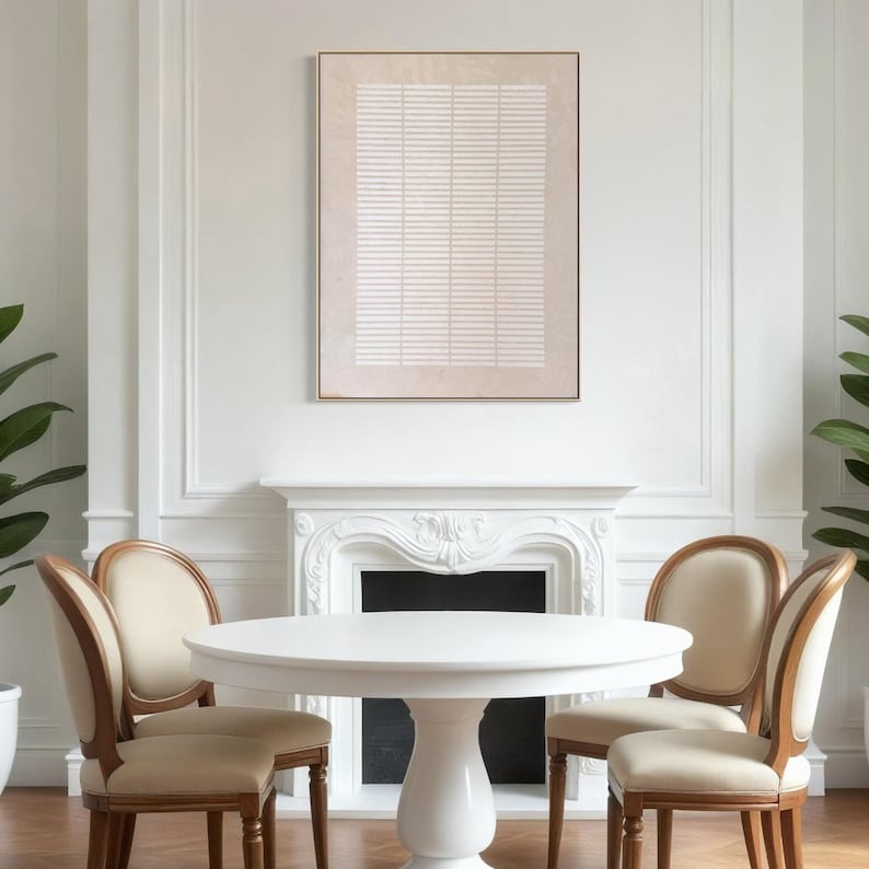 May include: A dining room scene featuring a white round table and four chairs with beige upholstery and wooden frames. A framed abstract artwork with a beige background and white horizontal lines hangs above a white fireplace. The room has white walls and hardwood floors.