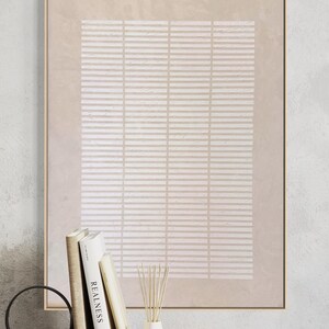 May include: A framed abstract art print with a beige and white geometric pattern. The artwork is displayed on a wooden surface with books, a decorative ring, and a small vase with reeds. The frame is gold.