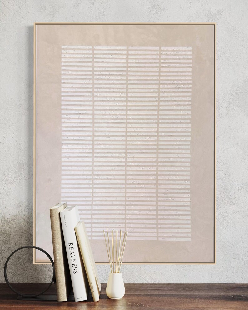 May include: A framed abstract art print with a beige and white geometric pattern. The artwork is displayed on a wooden surface with books, a decorative ring, and a small vase with reeds. The frame is gold.
