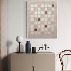 K&ouml;nnte beinhalten: Abstrakte Wandkunst mit einem Raster aus quadratischen Feldern in Beige- und Braunt&ouml;nen, gerahmt in hellem Holz. Eine moderne Innenszene zeigt eine Lampe, eine blaue Vase, einen schwarzen Zylinder und dekorative Objekte auf einem Schrank.