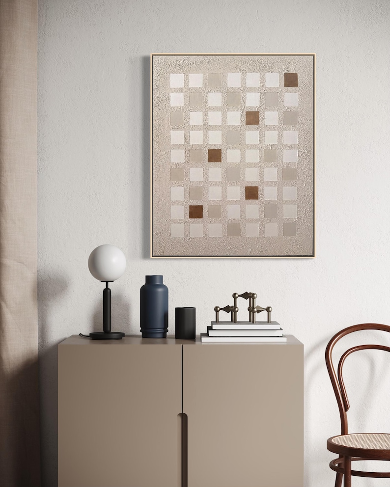 K&ouml;nnte beinhalten: Abstrakte Wandkunst mit einem Raster aus quadratischen Feldern in Beige- und Braunt&ouml;nen, gerahmt in hellem Holz. Eine moderne Innenszene zeigt eine Lampe, eine blaue Vase, einen schwarzen Zylinder und dekorative Objekte auf einem Schrank.