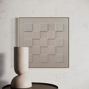 Pu&ograve; includere: Un'opera d'arte murale quadrata con un motivo a scacchiera beige monocromatico con quadrati testurizzati lisci e a coste alternati, incorniciata da un sottile bordo in legno chiaro, accanto a un vaso cilindrico alto di colore beige testurizzato.