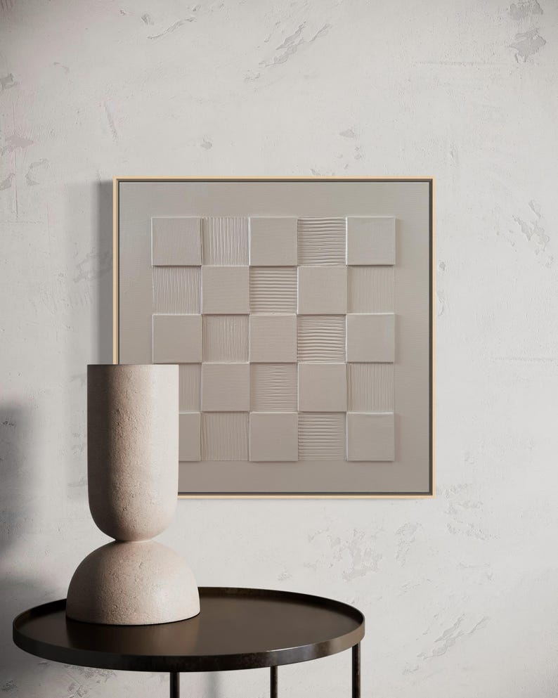 Pu&ograve; includere: Un'opera d'arte murale quadrata con un motivo a scacchiera beige monocromatico con quadrati testurizzati lisci e a coste alternati, incorniciata da un sottile bordo in legno chiaro, accanto a un vaso cilindrico alto di colore beige testurizzato.