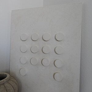 Peut inclure: Grande &oelig;uvre d'art murale rectangulaire blanc cass&eacute; avec une surface textur&eacute;e. Elle pr&eacute;sente une grille d'&eacute;l&eacute;ments circulaires en relief. Un vase beige textur&eacute; est visible en bas &agrave; gauche. L'&oelig;uvre est appuy&eacute;e contre un mur blanc.