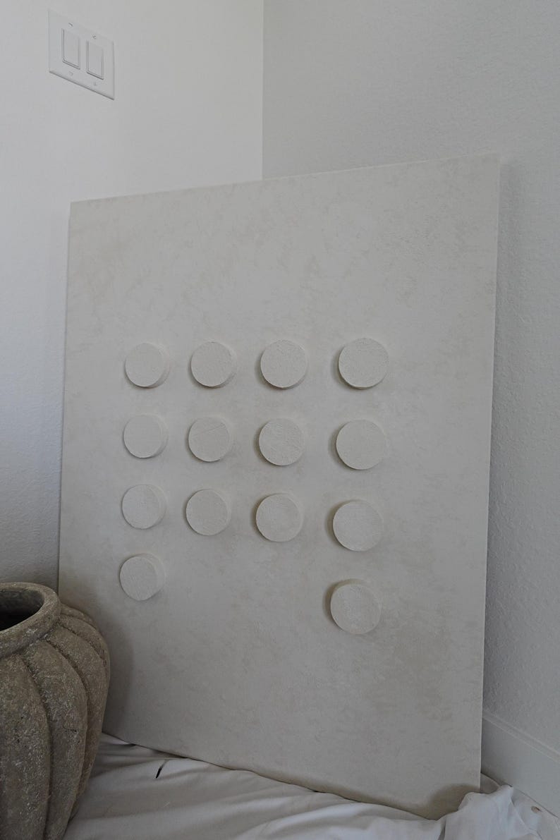 Peut inclure: Grande &oelig;uvre d'art murale rectangulaire blanc cass&eacute; avec une surface textur&eacute;e. Elle pr&eacute;sente une grille d'&eacute;l&eacute;ments circulaires en relief. Un vase beige textur&eacute; est visible en bas &agrave; gauche. L'&oelig;uvre est appuy&eacute;e contre un mur blanc.