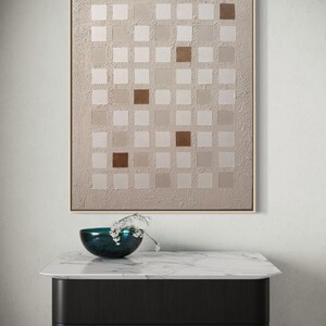 K&ouml;nnte beinhalten: Abstrakte Wandkunst mit einem Raster aus quadratischen Feldern in Beige- und Braunt&ouml;nen, goldgerahmt. Darunter eine dunkelblaue Schale mit wei&szlig;en Blumen auf einem Tisch mit Marmorplatte und schwarzen Beinen.