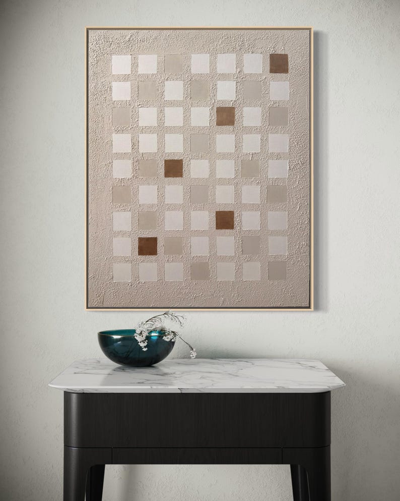 K&ouml;nnte beinhalten: Abstrakte Wandkunst mit einem Raster aus quadratischen Feldern in Beige- und Braunt&ouml;nen, goldgerahmt. Darunter eine dunkelblaue Schale mit wei&szlig;en Blumen auf einem Tisch mit Marmorplatte und schwarzen Beinen.