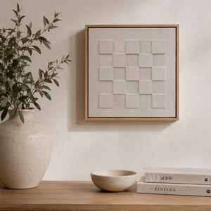Pu&ograve; includere: Un'opera d'arte da parete quadrata con un motivo a scacchiera beige chiaro, caratterizzata da quadrati in rilievo che alternano superfici lisce e texture a righe orizzontali, racchiusa in una sottile cornice in legno naturale.