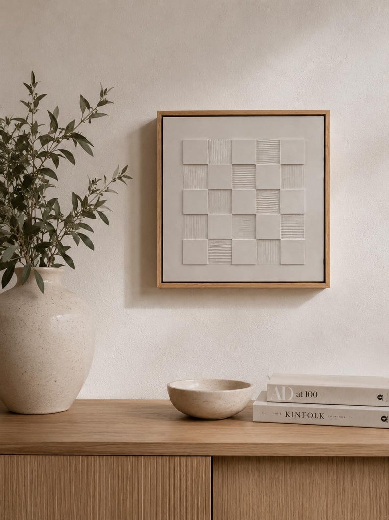 Pu&ograve; includere: Un'opera d'arte da parete quadrata con un motivo a scacchiera beige chiaro, caratterizzata da quadrati in rilievo che alternano superfici lisce e texture a righe orizzontali, racchiusa in una sottile cornice in legno naturale.
