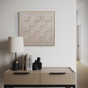 Pu&ograve; includere: Un'opera d'arte da parete quadrata caratterizzata da una griglia beige di quadrati in rilievo con superfici alternate lisce e testurizzate, incorniciata in legno chiaro. L'opera &egrave; esposta sopra una console beige con maniglie nere.