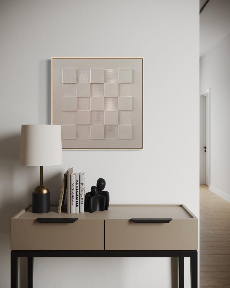 Pu&ograve; includere: Un'opera d'arte da parete quadrata caratterizzata da una griglia beige di quadrati in rilievo con superfici alternate lisce e testurizzate, incorniciata in legno chiaro. L'opera &egrave; esposta sopra una console beige con maniglie nere.