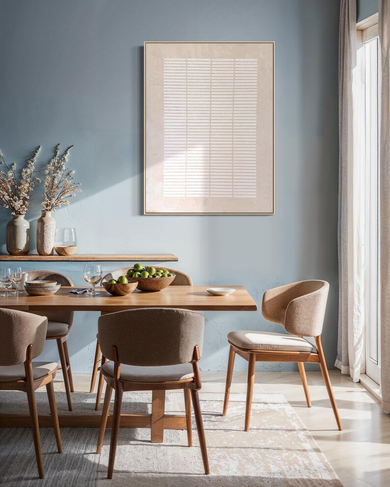 May include: A dining room scene featuring a light wood dining table and chairs. The chairs have curved backs and light-colored upholstery. A framed abstract artwork hangs on the blue wall. A wooden shelf holds vases and glassware. A bowl of limes sits on the table.