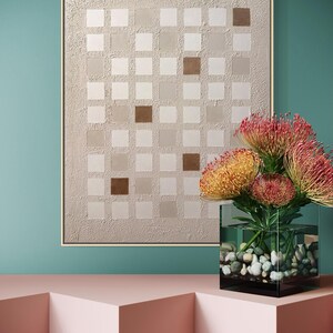 K&ouml;nnte beinhalten: Abstrakte Wandkunst mit einem Rastermuster aus Quadraten in Beige- und Braunt&ouml;nen, gerahmt in hellem Holz. Eine klare Glasvase mit bunten Blumen und Kieselsteinen steht auf einer rosa Oberfl&auml;che vor einer t&uuml;rkisfarbenen Wand.