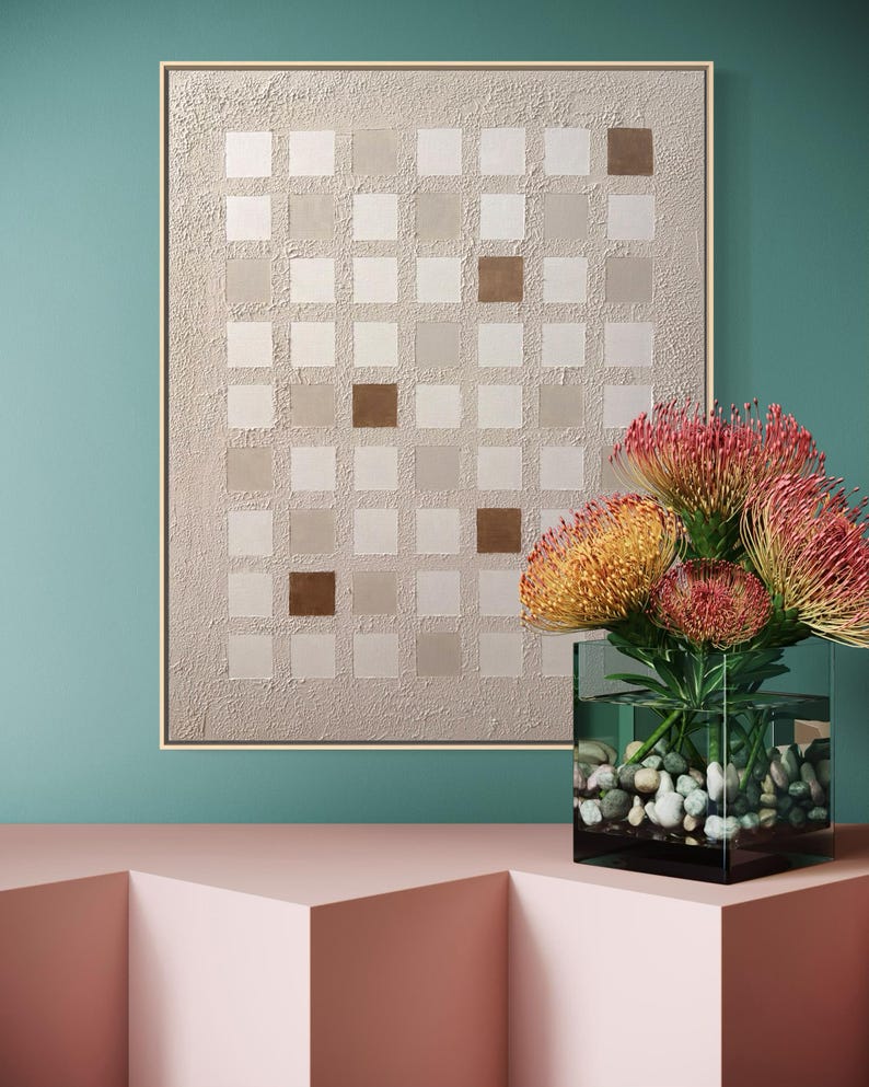 K&ouml;nnte beinhalten: Abstrakte Wandkunst mit einem Rastermuster aus Quadraten in Beige- und Braunt&ouml;nen, gerahmt in hellem Holz. Eine klare Glasvase mit bunten Blumen und Kieselsteinen steht auf einer rosa Oberfl&auml;che vor einer t&uuml;rkisfarbenen Wand.